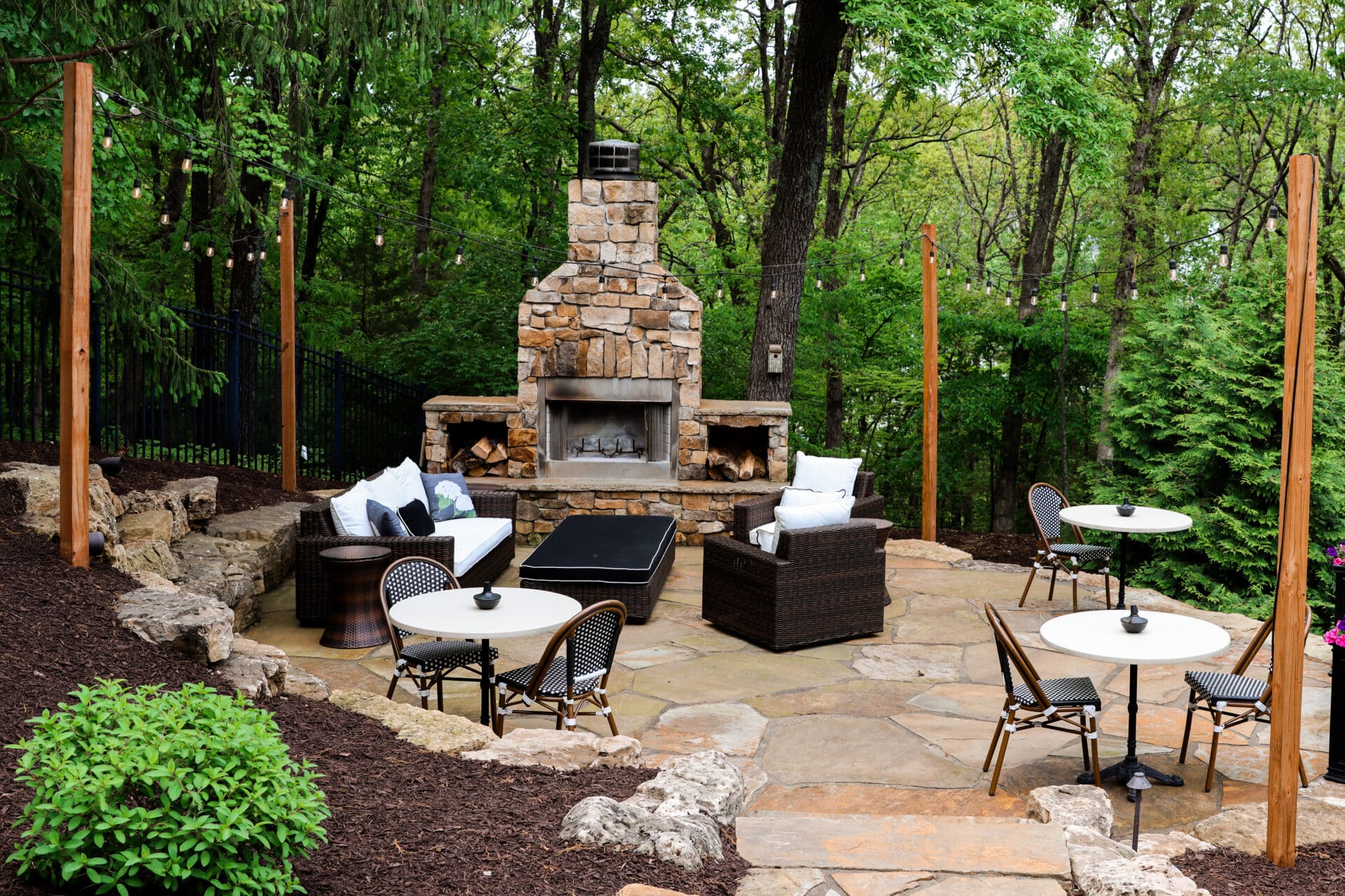 Cozy outdoor patio with stone fireplace, surrounded by lush greenery. Comfortable seating arrangements and string lights create a peaceful, inviting atmosphere.